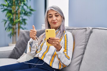 Wall Mural - Middle age woman with grey hair using smartphone sitting on the sofa smiling happy and positive, thumb up doing excellent and approval sign