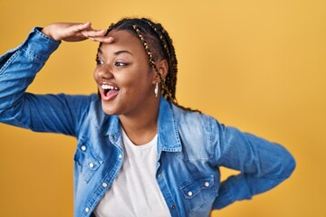 Wall Mural - African american woman with braids standing over yellow background very happy and smiling looking far away with hand over head. searching concept.