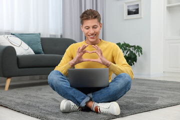 Wall Mural - Happy young man having video chat via laptop and making heart with hands on carpet indoors. Long-distance relationship