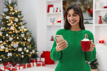 Wall Mural - Smiling woman with cup of Christmas cocoa and smartphone at home. Space for text