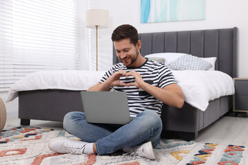 Canvas Print - Happy man making heart with hands during video chat via laptop in bedroom. Long-distance relationship