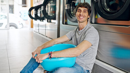 Poster - Young hispanic man sitting on floor leaning on washing machine at laundry facility