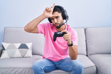 Canvas Print - Hispanic young man playing video game holding controller sitting on the sofa making fun of people with fingers on forehead doing loser gesture mocking and insulting.