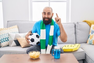 Canvas Print - Young hispanic man with beard and tattoos football hooligan holding ball supporting team surprised with an idea or question pointing finger with happy face, number one