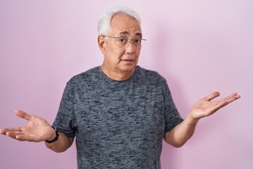 Sticker - Middle age man with grey hair standing over pink background clueless and confused expression with arms and hands raised. doubt concept.