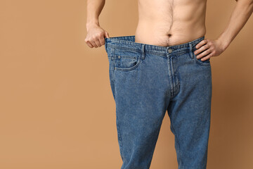 Wall Mural - Handsome sporty young man in loose jeans on brown background, closeup. Weight loss concept