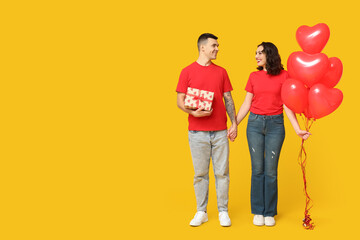 Poster - Loving young couple with heart-shaped balloons and gift box on yellow background. Celebration of Saint Valentine's Day