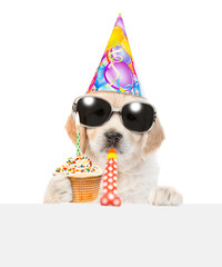 Poster - Golden retriever puppy wearing  sunglasses and party cap blows into party horn, looks above empty white banner and shows cupcake with burning candle. isolated on white background