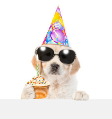 Poster - Golden retriever puppy wearing sunglasses and party cap looks above empty white banner and holds cupcake with burning candle. isolated on white background