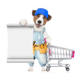 Poster - Funny Jack russell terrier puppy wearing red cap and denim overalls with tool belt shows empty list and holds the shopping cart. isolated on white background