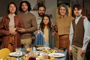 Family of six members of various generations standing by served table with burning menorah candles and homemade food and looking at you