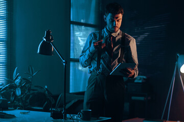 Poster - Young man in retro formalwear looking at screen of tablet while standing by his workplace in dark office and scrolling through online data