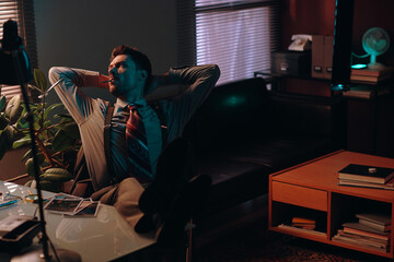 Aufkleber - Young relaxed man in formalwear keeping hands on back of head while sitting by table in dark office and smoking cigarette