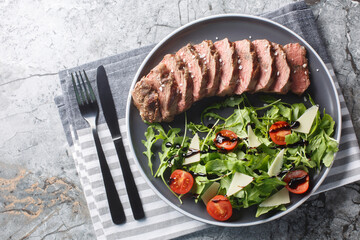 Traditional Italian main dish Beef tagliata tender steak, arugula, cherry tomatoes, Grana Padano shavings, balsamic reduction close-up on a plate on the table. Horizontal top view from above