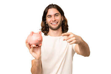 Wall Mural - Young handsome man holding a piggybank over isolated background points finger at you with a confident expression