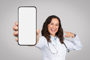 Excited woman doctor showing smartphone with blank screen at camera while standing over grey background, mockup