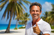 handsome middle aged man standing on a Caribbean beach with palm trees showing the victory sign