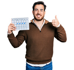 Wall Mural - Young hispanic man holding travel calendar smiling happy and positive, thumb up doing excellent and approval sign