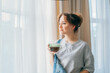 © okrasiuk - Free Time To Relax. Young woman looking out the window while overlooking the city at cozy home holding cup of hot tea drink. Happy calm female taking break for mental health wellbeing. No stress.