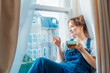 Free Time To Relax. Young woman looks out the window overlooking the city, sits on the windowsill at cozy home holds cup of hot tea drink. Happy calm female taking break for mental health wellbeing