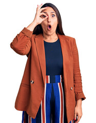 Sticker - Young beautiful brunette woman wearing elegant clothes doing ok gesture shocked with surprised face, eye looking through fingers. unbelieving expression.