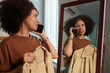 © DragonImages - Cheerful woman standing at mirror with shirt on hanger and talking on phone with friend