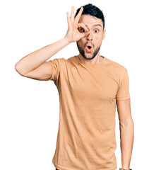 Canvas Print - Hispanic man with beard wearing casual t shirt doing ok gesture shocked with surprised face, eye looking through fingers. unbelieving expression.