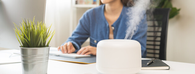 Modern air humidifier device during sitting workplace, happy asian young woman using computer work on internet, enjoying aromatherapy steam scent from essential oil diffuser in room at home office.