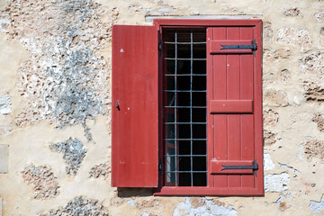 Wall Mural - Window with wooden red shutter and metal grid on peeled wall background. Greek architecture.