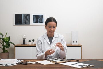 Poster - Virtual Prescription: During an online call with a patient, a female doctor holds a bottle of pills or vitamins on her desk, conducting telehealth consulting and prescribing medication. 