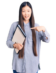 Sticker - Young beautiful chinese woman holding notebook smiling happy pointing with hand and finger