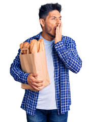 Wall Mural - Handsome latin american young man holding paper bag with bread bored yawning tired covering mouth with hand. restless and sleepiness.