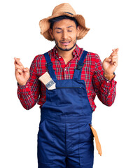 Wall Mural - Handsome latin american young man weaing handyman uniform gesturing finger crossed smiling with hope and eyes closed. luck and superstitious concept.