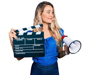 Poster - Young blonde woman holding video film clapboard and megaphone smiling looking to the side and staring away thinking.