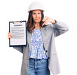 Young beautiful brunette woman wearing architect hardhat holding contract with angry face, negative sign showing dislike with thumbs down, rejection concept
