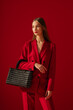 Fashionable confident woman wearing trendy red suit blazer, classic trousers, holding stylish black tweed bag, handbag, posing on red background. Studio fashion portrait. Copy, empty space for text
