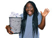 Young black woman with braids holding paper bin full of crumpled papers celebrating victory with happy smile and winner expression with raised hands