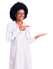 Poster - Young african american woman wearing doctor coat and stethoscope amazed and smiling to the camera while presenting with hand and pointing with finger.