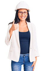 Poster - Young beautiful latin girl wearing architect hardhat and glasses doing happy thumbs up gesture with hand. approving expression looking at the camera showing success.