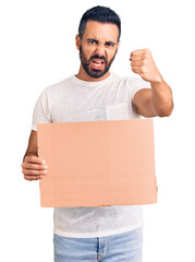 Poster - Young hispanic man holding banner cardboard annoyed and frustrated shouting with anger, yelling crazy with anger and hand raised