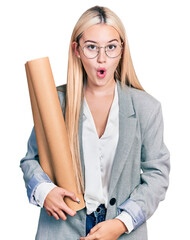 Wall Mural - Beautiful blonde woman holding paper blueprints scared and amazed with open mouth for surprise, disbelief face