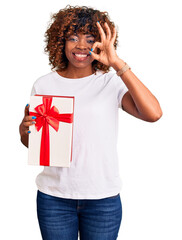 Poster - Young african american woman holding gift doing ok sign with fingers, smiling friendly gesturing excellent symbol