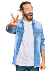 Wall Mural - Attractive man with long hair and beard wearing casual denim jacket smiling with happy face winking at the camera doing victory sign with fingers. number two.