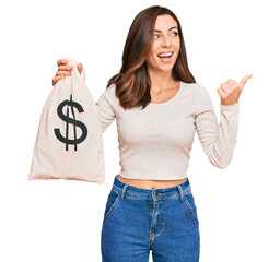 Canvas Print - Young brunette woman holding dollars bag pointing thumb up to the side smiling happy with open mouth