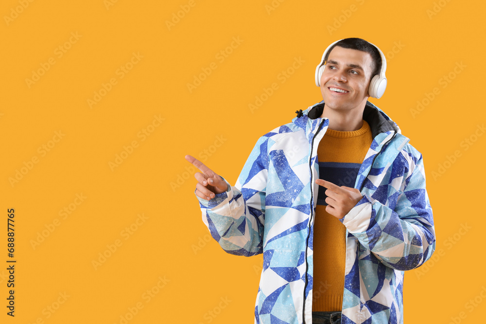 Young man in warm jacket with headphones pointing at something on yellow background