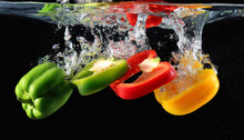 Slices Of Ripe Red, Green And Yellow Pepper Falling Into The Water With A Splash On Black Background