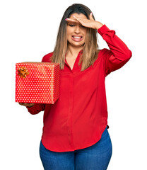 Poster - Beautiful brunette woman holding gift stressed and frustrated with hand on head, surprised and angry face