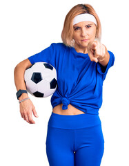 Sticker - Young blonde woman holding soccer ball pointing with finger to the camera and to you, confident gesture looking serious