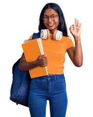 Wall Mural - Young indian girl holding student backpack and books doing ok sign with fingers, smiling friendly gesturing excellent symbol