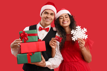 Sticker - Happy young couple in Santa hats with gift card, present boxes and decorative snowflake on red background. Christmas shopping concept